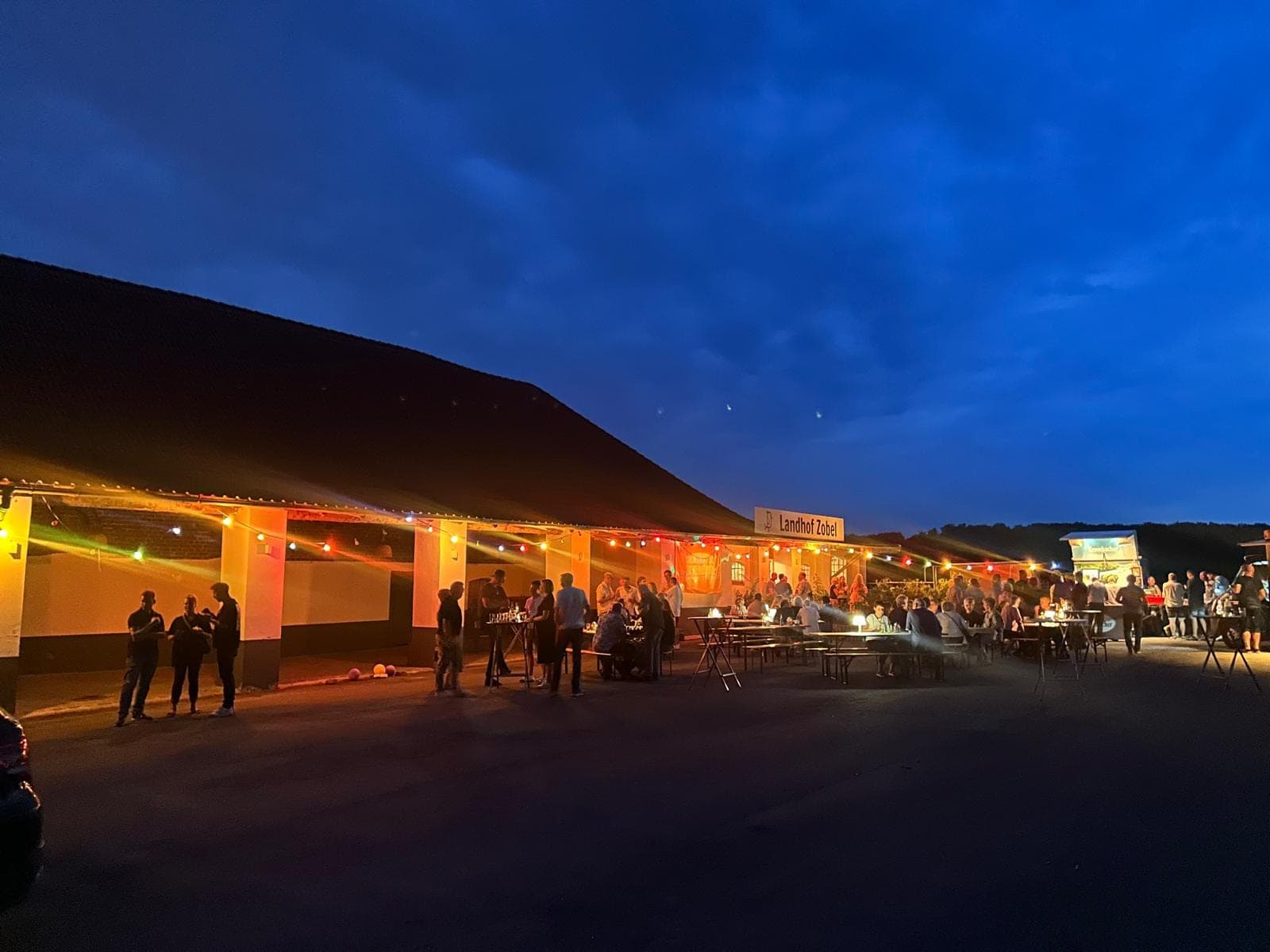 Abendliche Szene mit beleuchteter Terrasse, Menschen sitzen an Tischen und stehen vor einem Gebäude.