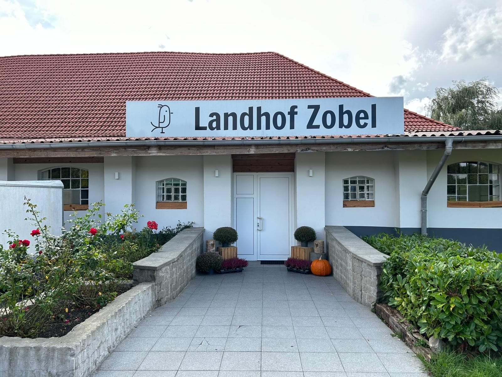 Eingang zum Landhof Zobel mit weißer Tür, roten Dachziegeln und Bepflanzung.