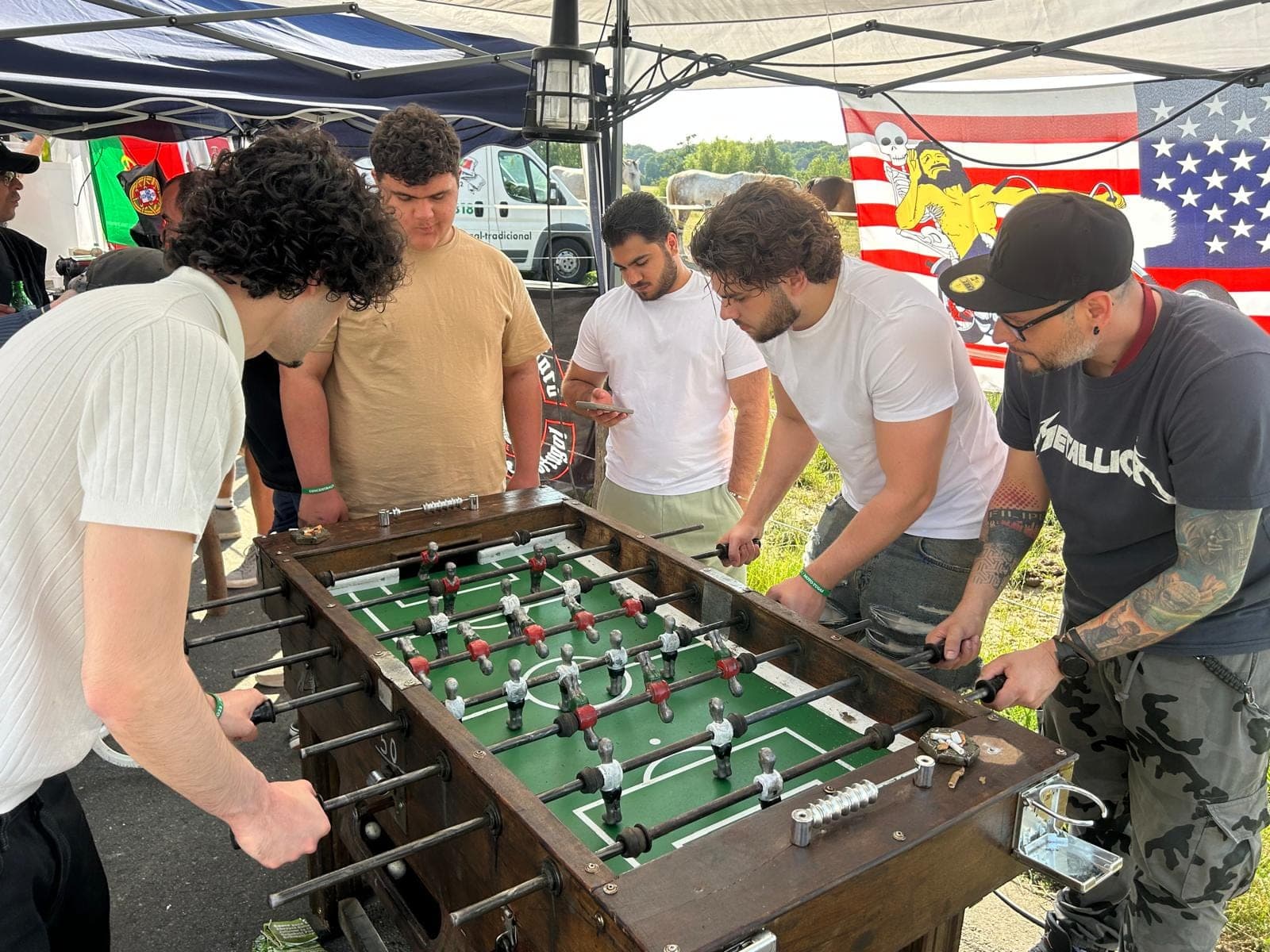 Fünf junge Männer spielen unter einem Zelt Tischfußball; eine US-Flagge hängt im Hintergrund.