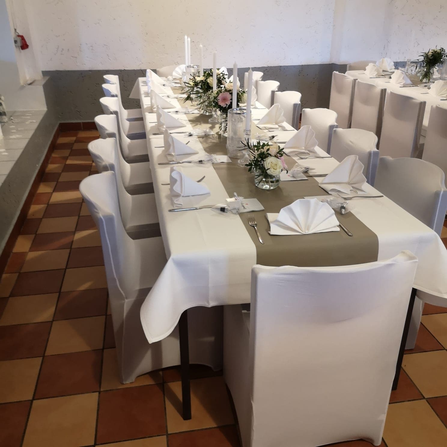 Lange, festlich gedeckte Tische mit weißen Hussen und grauen Läufern, dekoriert mit Blumen und Kerzen.