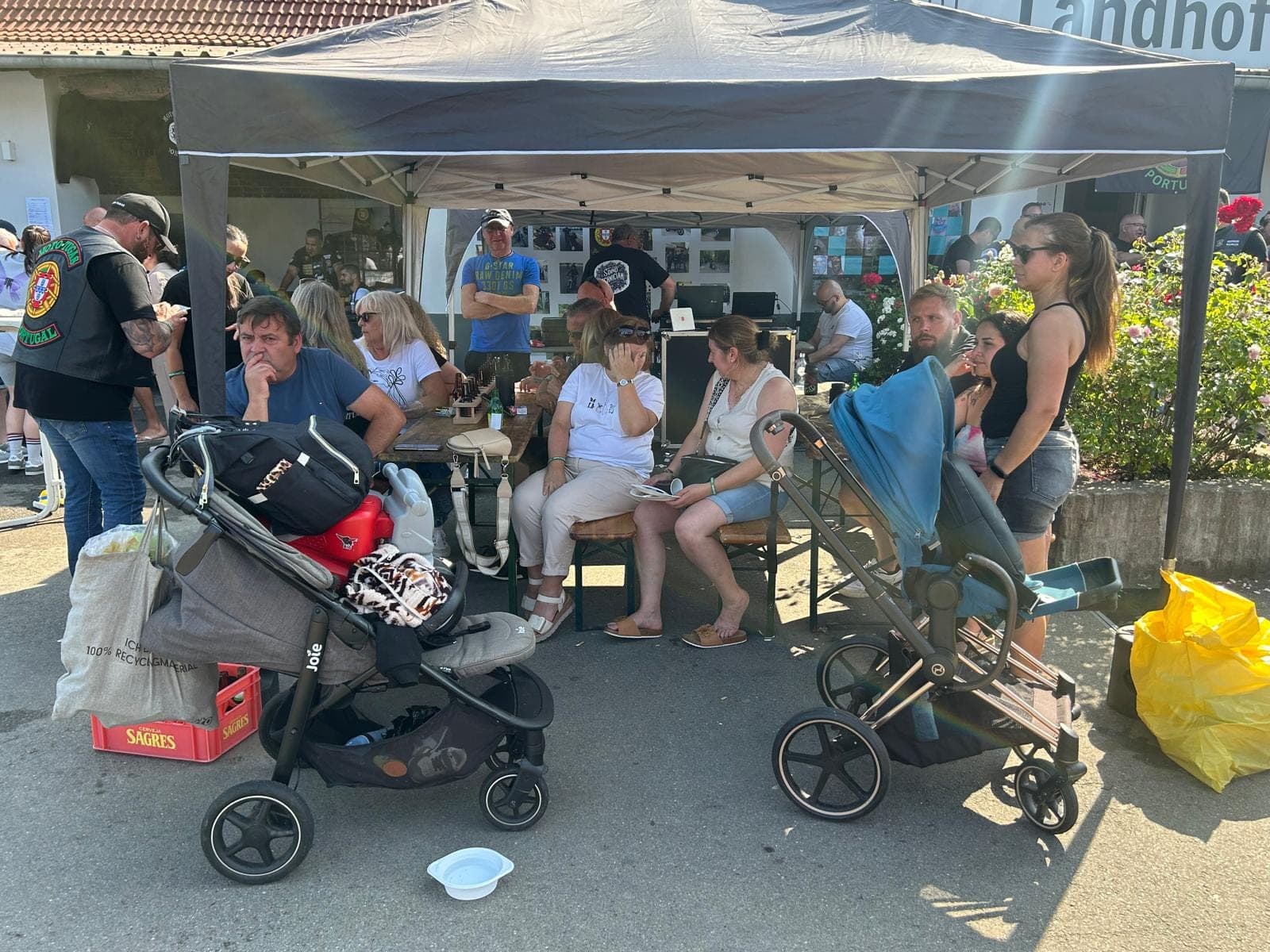Menschen sitzen unter einem Zelt, zwei Kinderwagen stehen im Vordergrund.