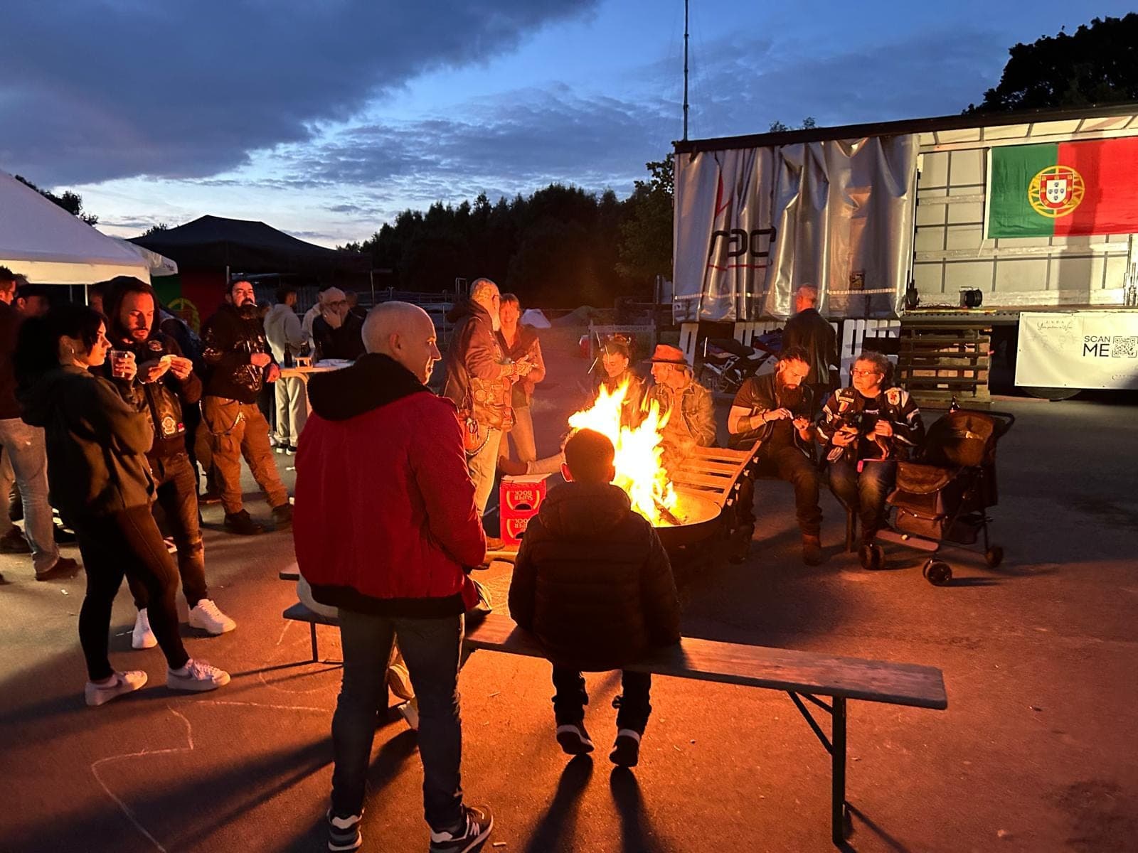 Menschen versammeln sich um ein Lagerfeuer vor einem Anhänger mit portugiesischer Flagge in der Abenddämmerung.