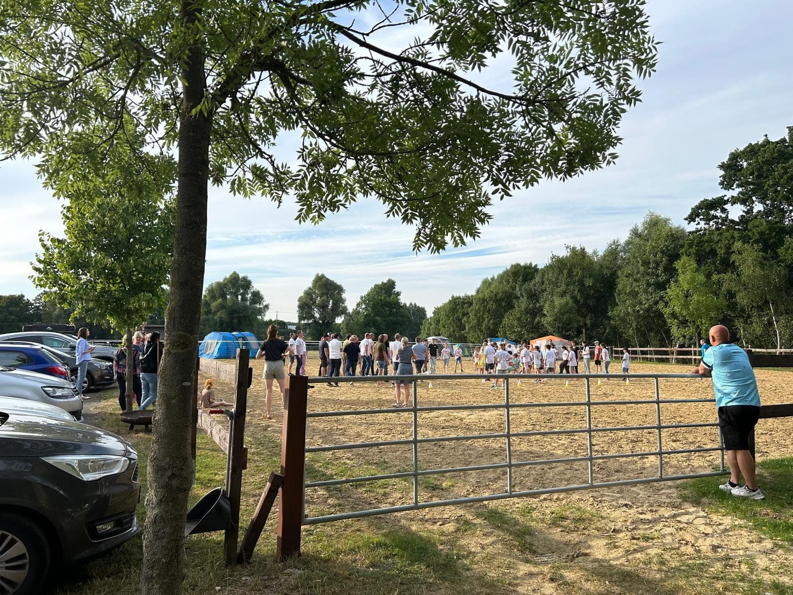 Menschengruppe auf einem Sportfeld, umgeben von einem Holzzaun; Autos parken links unter einem Baum.