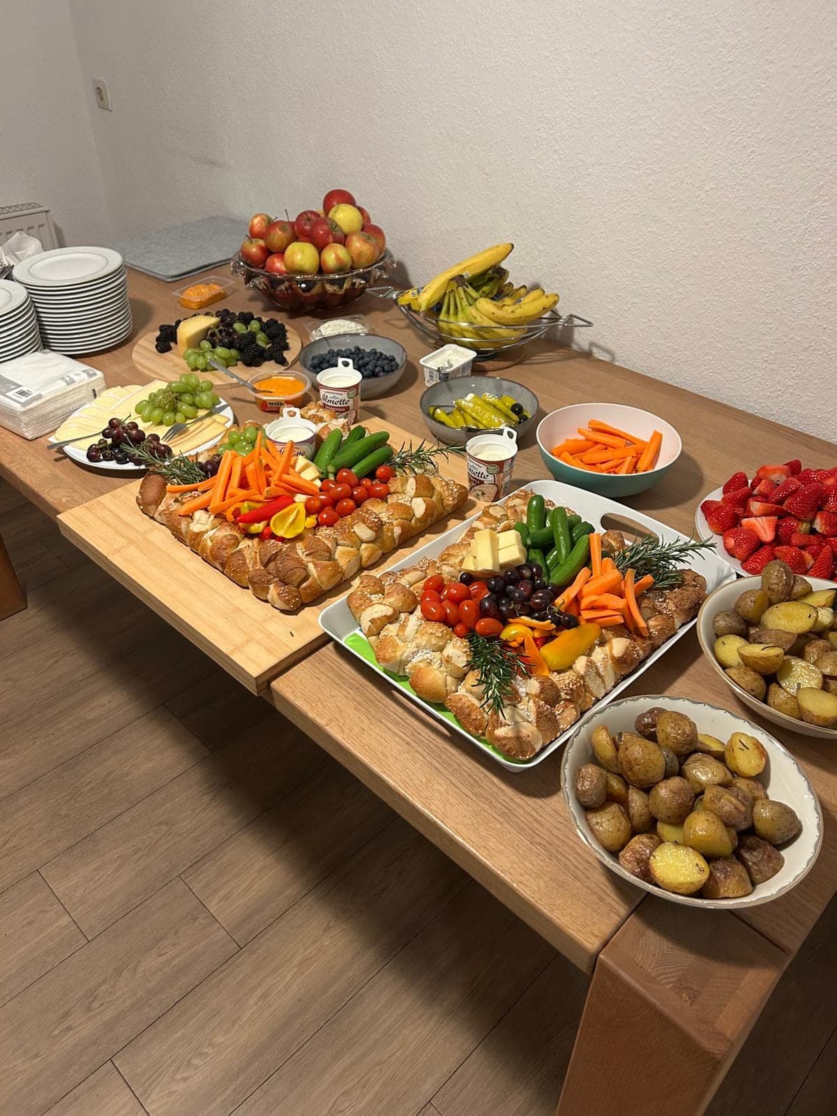 Ein Tisch mit verschiedenen Speisen, darunter zwei Brotzöpfe mit Gemüse, Obst, Käse und Kartoffeln.